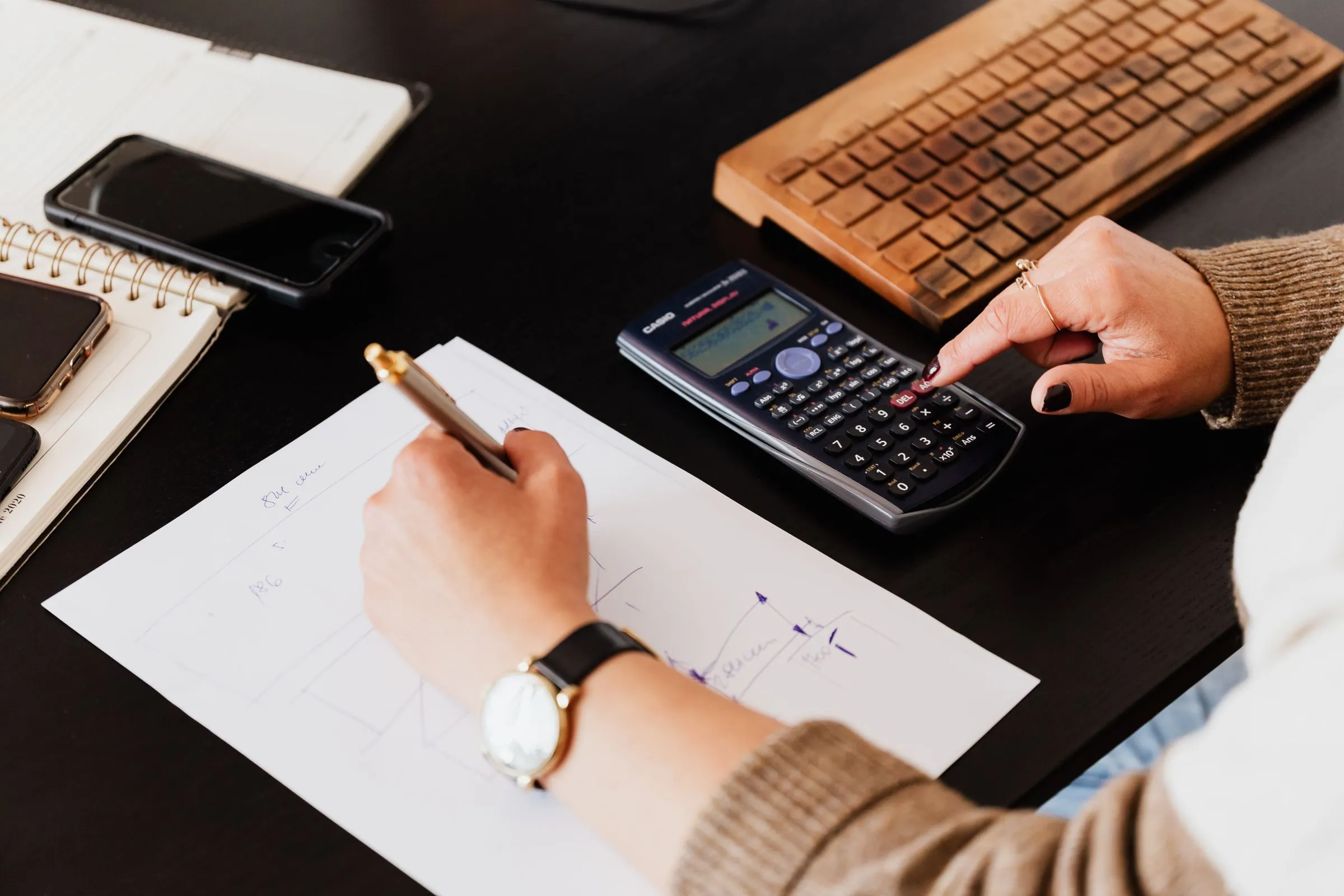Mãos fazendo contas com calculadora e anotando numa folha sobre a mesa